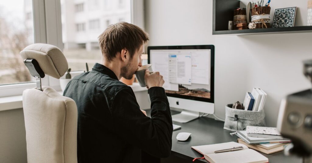 Person working at a desk, drinking coffee while using a computer at home.