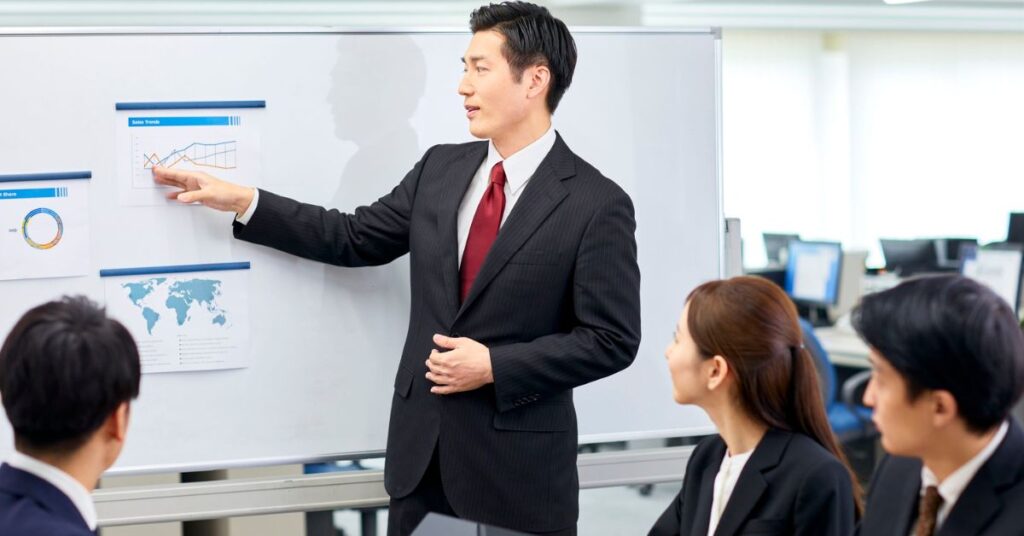 Business professional presenting charts on a whiteboard to colleagues in a meeting.