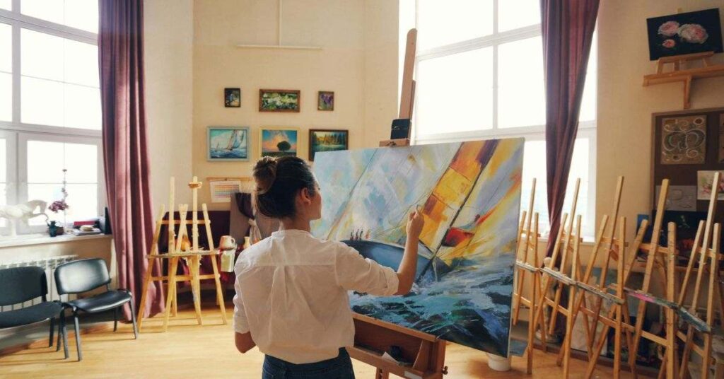 Artist painting a colorful sailboat on canvas in a bright art studio.