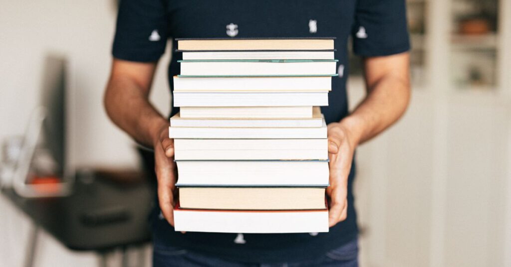 Person holding a large stack of books in hands, symbolizing education, study, and knowledge growth concept.